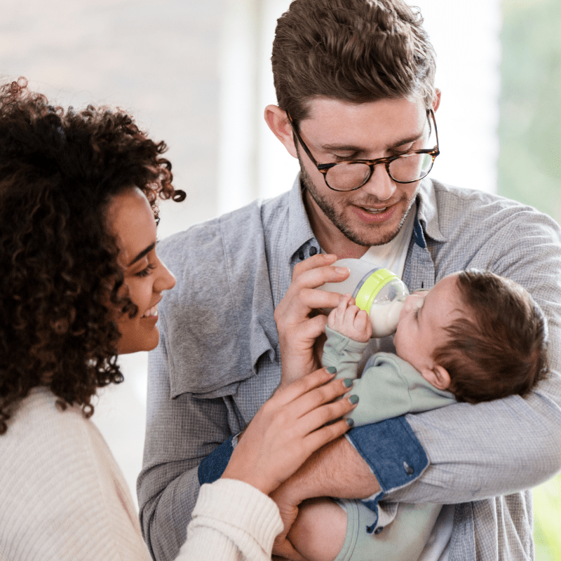 A new father holds his infant child and feeds her with a bottle as his wife looks on.